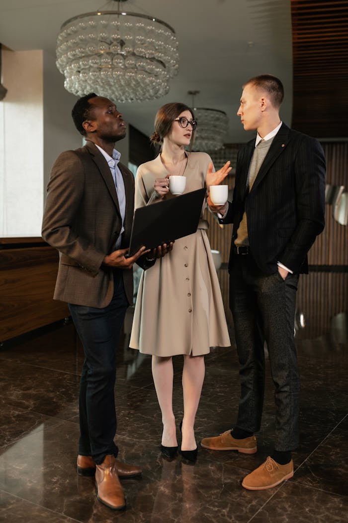 Three professionals engaged in a business discussion, holding coffee cups and a laptop, in a modern office setting.